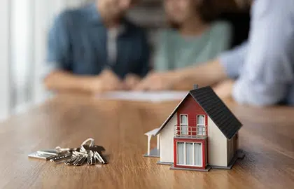 A model house and keys on a desk in focus, and an out of focus couple talking with a mortgage advisor in the background